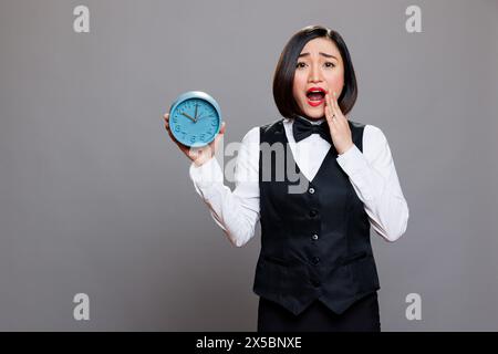 Ängstliche asiatische Kellnerin, die Panik hat, während sie den Wecker mit dem Kreiskalender hält und mit offenem Mund in die Kamera blickt. Junge verängstigte Frau an der Rezeption zeigt ein Zeituhr-Porträt Stockfoto