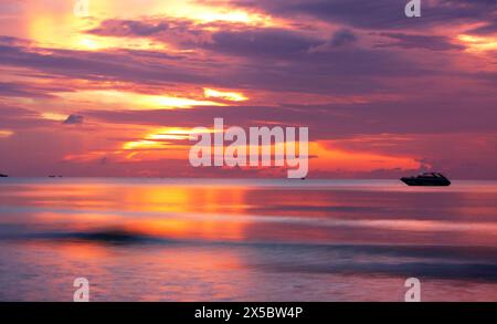 Sonnenaufgang am Chaweng Beach, Koh Samui, mit Blick auf das Meer, die Sonne teilweise von Wolken versteckt, reflektiert vom Wasser, mit dem Boot eine Silhouette. Stockfoto