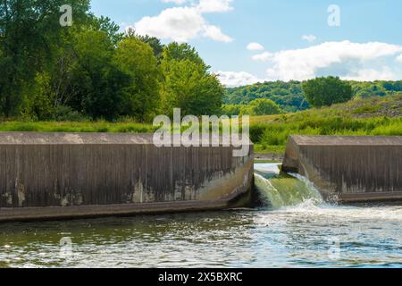 Das Wasser fließt durch das Schleusentor in einem Betondamm auf einem Fluss, mit grünen Bäumen und Vegetation auf den Hügeln im Hintergrund. Stockfoto