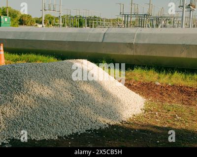 Weite Sicht über grünes Gras, weiße Steine anzuhäufen und auf dem Boden einen Versorgungsmastabschnitt aus Stahl mit einer elektrischen Unterstation hinten zu legen. Handfläche Stockfoto