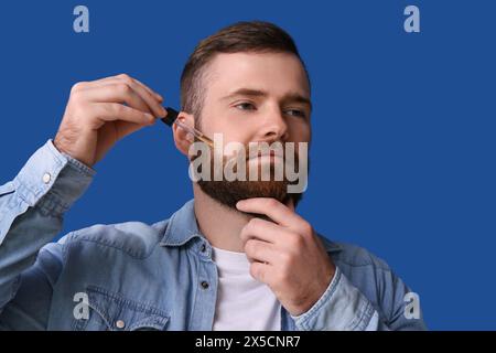 Junger bärtiger Mann, der ätherisches Öl auf seinem Gesicht vor blauem Hintergrund aufträgt Stockfoto