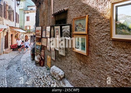 Gardasee, Italien, 16. April 2024: Enge Gasse mit runden Kopfsteinpflastern und Kunstwerken, die an Touristen in einem Haus im Dorf Malcesine verkauft werden Stockfoto