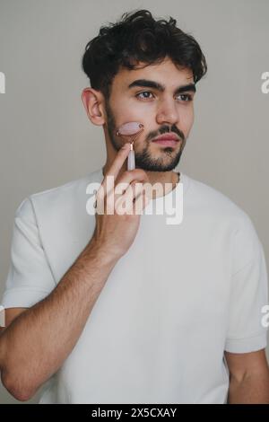 Junger brünette Mann mit weißem T-Shirt, der wegschaut und sein Gesicht mit Jadewalze massiert. Körper- und Hautpflege. Isoliert auf weißem Hintergrund. Studio Stockfoto