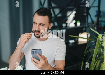 Porträt eines kaukasischen Mannes, der ein weißes T-Shirt mit Bart mit Jaderolle trägt, während er das Gesicht im Stehen zu Hause massiert. Hautbehandlung. Schönheitsprodukt. Stockfoto