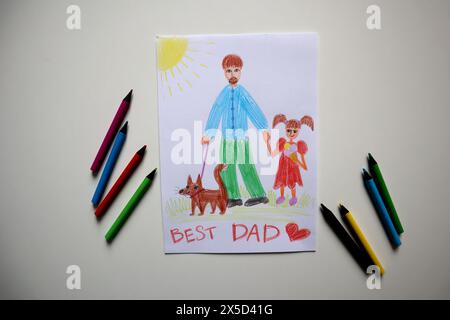 Vatertagskarte von Kind gezeichnet. Weißer Hintergrund. Buntstifte in der Nähe der Postkarte. Die Karte zeigt den Vater, der mit seiner Tochter und seinem Hund läuft. Sie Stockfoto