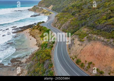 Aus der Vogelperspektive sehen Sie Wellen, die unter einer gewundenen Straße entlang einer dramatischen Küste in der Nähe von Lorne an der Great Ocean Road in Victoria, Australien, zusammenbrechen. Stockfoto