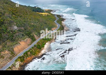 Aus der Vogelperspektive sehen Sie Wellen, die unter einer gewundenen Straße entlang einer dramatischen Küste in der Nähe von Lorne an der Great Ocean Road in Victoria, Australien, zusammenbrechen. Stockfoto