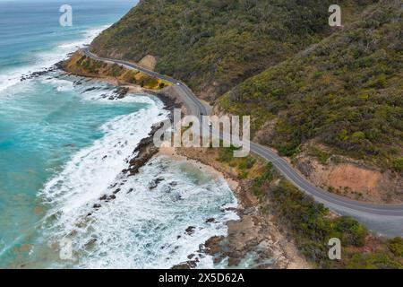 Aus der Vogelperspektive sehen Sie Wellen, die unter einer gewundenen Straße entlang einer dramatischen Küste in der Nähe von Lorne an der Great Ocean Road in Victoria, Australien, zusammenbrechen. Stockfoto