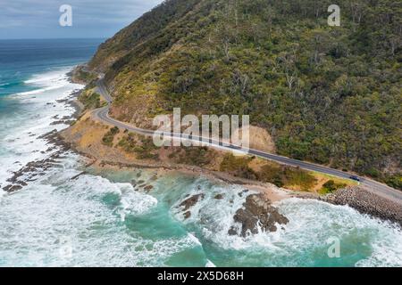 Aus der Vogelperspektive sehen Sie Wellen, die unter einer gewundenen Straße entlang einer dramatischen Küste in der Nähe von Lorne an der Great Ocean Road in Victoria, Australien, zusammenbrechen. Stockfoto