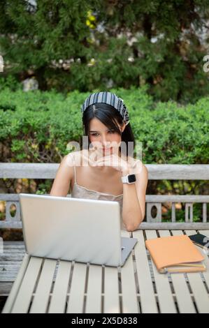 Eine zweifelhafte, verwirrte asiatische Frau, die an ihrem Laptop an einem Tisch in einem grünen Garten arbeitet, mit einer niedrigen Internetverbindung und einer funktionierenden Fernbedienung zu kämpfen hat Stockfoto