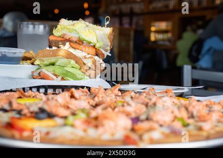 Lachsburger und unscharfe Lachspizza auf einem Tisch. Nicht erkennbare Leute im Restaurant. Alaska. Stockfoto