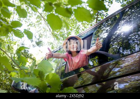 Ein kleiner Junge gibt die Daumen aus dem Fenster eines Autos hoch. Das Auto steht in einem Wald, und der Junge lächelt Stockfoto