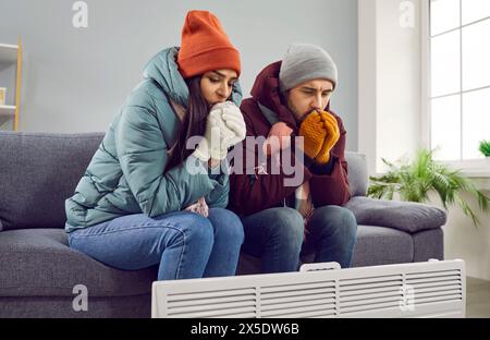 Junge attraktive Paare sitzen auf dem Sofa zu Hause in warmen Kleidern und wärmen sich die Hände mit Heizung im Wohnzimmer, frieren vor der Kälte Stockfoto
