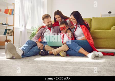 Glückliche Mutter, Vater und Kinder in roten Superheldenmänteln mit Laptop-Computer zusammen Stockfoto