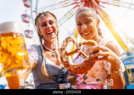 Freundinnen, die mit Brezel oder Brezen und Bierkrug auf einem bayerischen Jahrmarkt oder auf einem oktoberfest toben oder in Nationalkostüme oder Dirndl stummeln Stockfoto