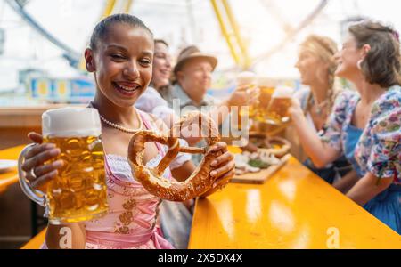 Eine junge Frau, die fröhlich einen Bierkrug und eine Brezel in einem Bierzelt hält, auf dem Oktoberfest oder der Dult in deutschland Stockfoto