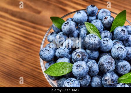 Heidelbeeren mit grünen Blättern in einer Glasplatte und Wassertropfen auf einem hölzernen Hintergrund Stockfoto