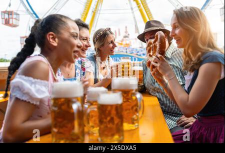 Freunde lachen und trinken Bier und Brezeln an einem Festzelttisch, lustige Fahrgeschäfte im Hintergrund auf dem münchner oktoberfest oder lauschen Stockfoto