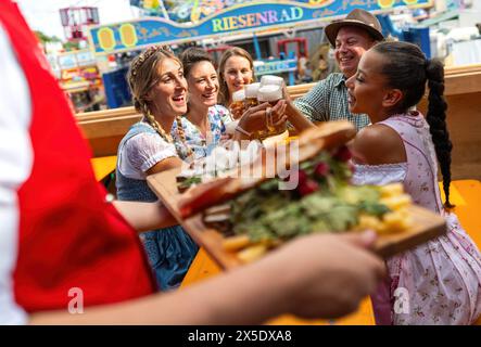 Freunde am oktoberfest Festtisch erhalten ein Tablett mit traditionellem Essen, toasten mit Bier, Vergnügungsfahrten im Hintergrund in münchen g Stockfoto