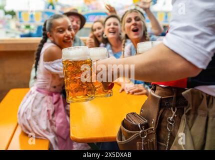 Kellner servieren Bier für eine fröhliche Gruppe von Freunden beim oktoberfest oder Duld in münchen, Vergnügungsfahrten im Hintergrund Stockfoto