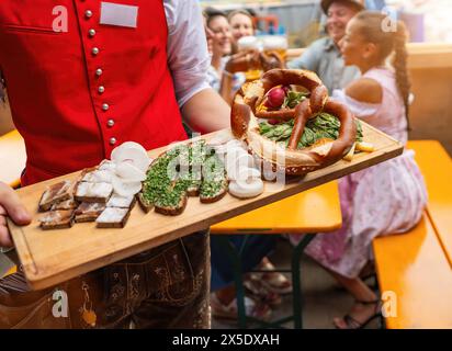 Kellner in roter Weste serviert traditionelle deutsche Gerichte auf einer Holzplatte zu einer Gruppe von Leuten in einem Bierkelzelt bei einem oktoberfest oder Dult auf deutsch Stockfoto