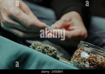 Nahaufnahme eines Mannes, der einen Joint rollt und an einem Tisch sitzt, wo Cannabisknospen und ein Glas mit Tabak sind Stockfoto
