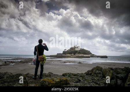 Ein Mann fotografiert St. Michael's Mount in Cornwall mit seinem Smartphone. Erinnert an das Gemälde der Wanderer über dem Nebelmeer von Caspar David Friedric Stockfoto