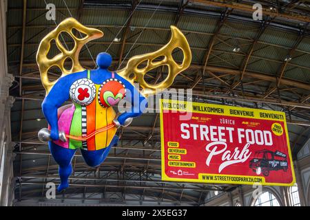 Zürich, Schweiz - 26. April 2024: Eine gigantische Nana in Form eines Schutzengels im Bahnhof Zürich und ein Street Food Festival Banner Stockfoto