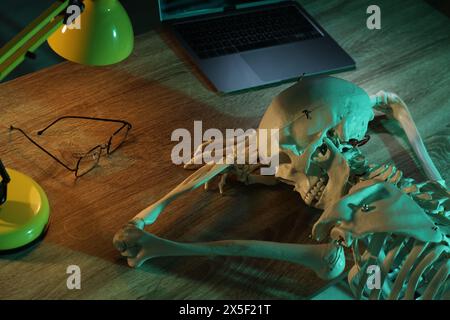 Wartendes Konzept. Menschliches Skelett, das an einem Holztisch mit Laptop schläft Stockfoto