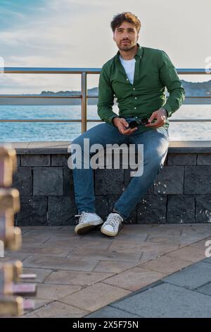 Ein junger Mann in grünem Hemd und Jeans sitzt lässig am Meer, hält eine Sonnenbrille und genießt den ruhigen Blick aufs Wasser. Stockfoto