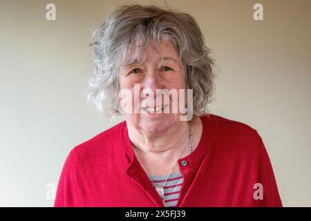 Eine reife Frau mit grauem Haar, die in einer dunkelroten Strickjacke steht und meist mit ernstem Ausdruck in die Kamera blickt Stockfoto