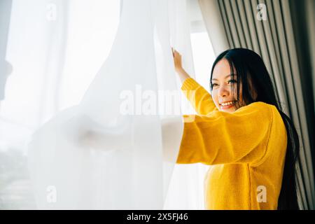 Eine junge Frau öffnet am frühen Morgen die Vorhänge und lächelt über die Aussicht und fühlt sich entspannt Stockfoto