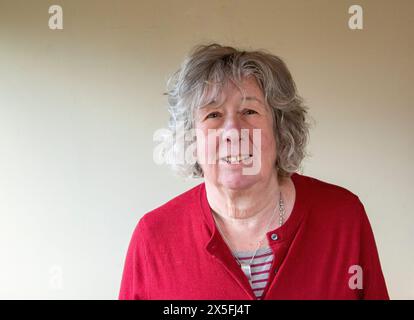 Eine reife Frau mit grauem Haar, die in einer dunkelroten Strickjacke steht und meist mit ernstem Ausdruck in die Kamera blickt Stockfoto