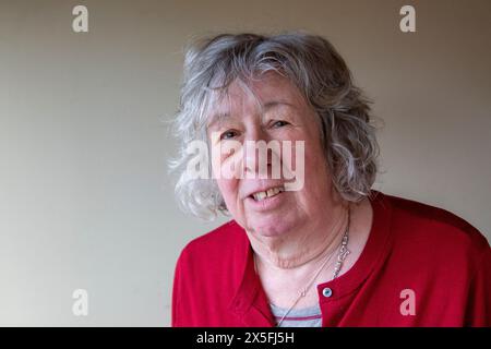 Eine reife Frau mit grauem Haar, die in einer dunkelroten Strickjacke steht und meist mit ernstem Ausdruck in die Kamera blickt Stockfoto