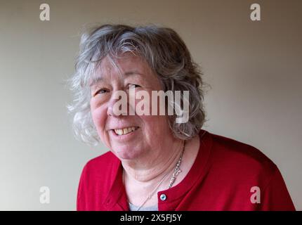 Eine reife Frau mit grauem Haar, die in einer dunkelroten Strickjacke steht und meist mit ernstem Ausdruck in die Kamera blickt Stockfoto