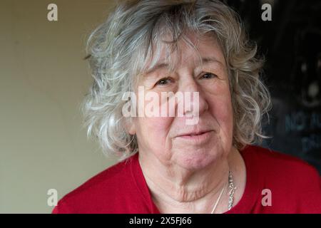 Eine reife Frau mit grauem Haar, die in einer dunkelroten Strickjacke steht und meist mit ernstem Ausdruck in die Kamera blickt Stockfoto