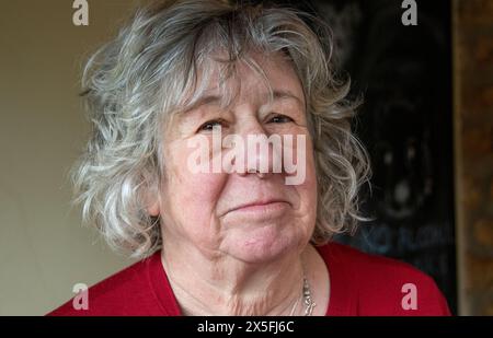 Eine reife Frau mit grauem Haar, die in einer dunkelroten Strickjacke steht und meist mit ernstem Ausdruck in die Kamera blickt Stockfoto