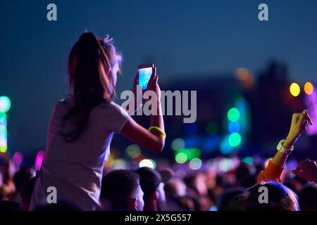 Girl filmt Musikkonzert auf Smartphone, inmitten lebendiger Festivalbesucher in der Dämmerung. Das Publikum genießt Live-Auftritte, Erinnerungen und lebendige Bühnenlichter Stockfoto