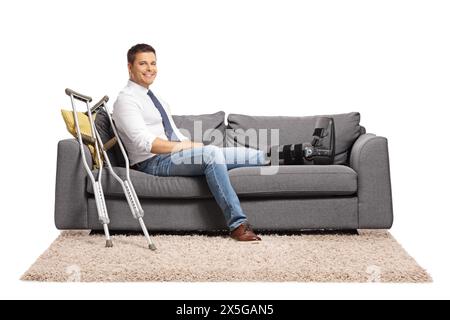 Geschäftsmann mit einer Fußverletzung und Beinstütze, der auf einem Sofa sitzt, isoliert auf weißem Hintergrund Stockfoto