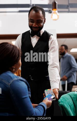 Kasselloses Hotel. Afroamerikanische Frau, die kontaktlos mit Kreditkarte bezahlt, während sie sich in der Lobby ausruht. Lächelnder Kellner, der das Terminal hält und Zahlungen über nfc-Technologie akzeptiert Stockfoto