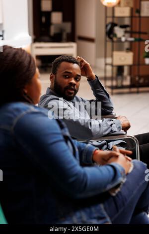 Junger afroamerikanischer Rollstuhlfahrer, der mit Begleitung reist, behinderter Tourist, der in der Hotellobby sitzt und mit Freundin oder Frau spricht, während er auf den Check-in wartet, Reisende mit Behinderungen Stockfoto