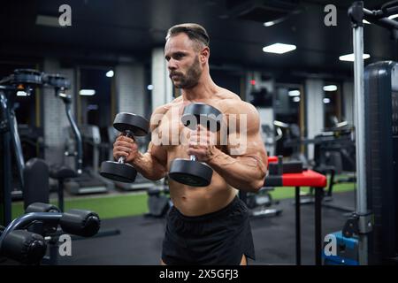 Entschlossener, gutaussehender männlicher Gewichtheber, der im Fitnessstudio arbeitet. Porträt eines bärtigen Mannes mit Frisur, Hanteln heben und beiseite schauen, vor verschwommenem Hintergrund. Bodybuilding, Sportkonzept. Stockfoto