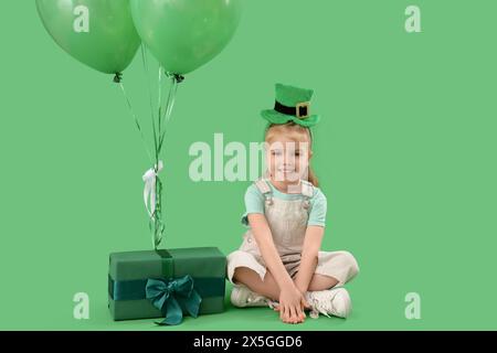 Niedliches kleines Mädchen mit Leprechaun Hut, Geschenkbox und Ballons auf grünem Hintergrund. St. Patrick's Day-Feier Stockfoto