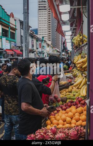Ein belebter Straßenmarkt erfüllt den Rahmen, bei dem verschiedene Käufer aus einer Auswahl an frischem Obst wie Bananen, Äpfel und Orangen wählen. Dieses l Stockfoto