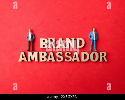 Miniatur-Leute mit Blick von oben und Holzwort mit Text BRAND AMBASSADOR auf rotem Hintergrund. Stockfoto