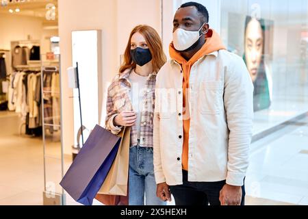 Pandemic Shopping. Attraktives Paar mit Schutzmaske medizinische Masken, die mit Einkaufstaschen in Mall Coronavirus Quarantäne, Kopierraum, schließen Stockfoto