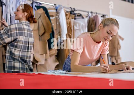 Selbstbewusster junger Modedesigner und kreativer Partner, der in einem neuen Projekt zusammen arbeitet und über eine neue Textilproduktion nachdenkt, wobei braune Kleidung verwendet wird. In einer modernen Werkstatt. Rothaarige Frau im Hintergrund Stockfoto