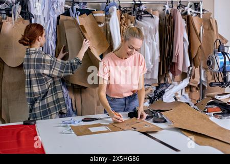 Junge erfolgreiche Modedesignerinnen, die in einer Textilfabrik arbeiten, Stoffe auswählen und Maßnahmen treffen. Zwei Arbeiterinnen, die in der Textilfabrik Kleidungsstücke zum Sticken vorbereiten Stockfoto