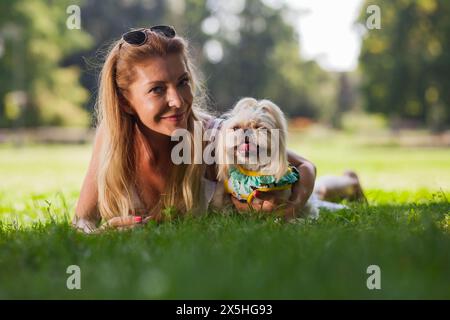 Fröhlicher Moment, in dem eine blonde Frau mit ihrem süßen Shih Tzu den Sommermonaten draußen genießt, beide entspannen auf dem grünen Gras in einer strahlenden Parklandschaft. Stockfoto