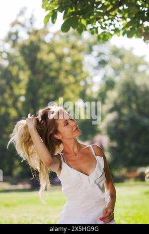 Eine fröhliche, blonde Frau in einem weißen Sommerkleid lächelt, während sie einen sonnigen Tag im Freien in einem üppig grünen Park genießt. Das Bild strahlt Glück und Freizeit aus. Stockfoto